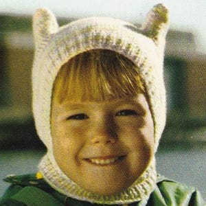 Puede incluir: Un niño con un gorro de punto color crema con dos pequeñas protuberancias en forma de orejas. El gorro forma parte de un conjunto que incluye un cuello a juego. El niño lleva una chaqueta verde.