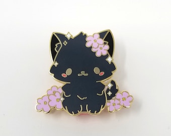 May include: Black enamel pin of a cute cartoon cat with pink flowers on its head and around its paws. The cat has a gold outline and a sparkly detail on its back.