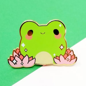 Puede incluir: Una rana verde con una flor rosa a cada lado, la rana tiene una expresión feliz y detalles brillantes.
