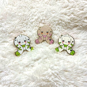 以下が含まれることがあります： 白い体、緑色の模様、ピンク色の耳を持つ漫画のキャラクターが描かれたエナメルピンが3つあります。キャラクターは腕を組んで座っています。ピンは白いふわふわの表面に置かれています。