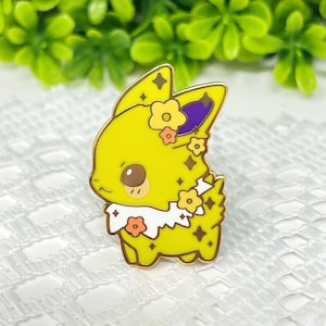 May include: Yellow enamel pin of a cartoon character with flowers and sparkles. The character has a white collar and purple ears.