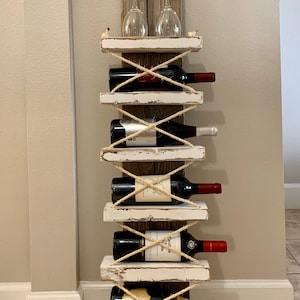 May include: White wooden wine rack with rope accents, holding six bottles of wine and two wine glasses. The rack has a distressed white paint finish and is mounted on a wall.