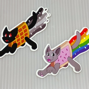 Puede incluir: Dos pegatinas de gato. Una es negra con un cuerpo de gofre y una estela gris oscuro con calaveras. La otra es gris con un cuerpo de Pop-Tart y una estela de arcoíris. Ambos gatos están en pose de carrera.