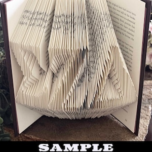 May include: A folded book art sculpture featuring the Greek letters Sigma, Zeta, and Delta created by precisely folding the individual pages of a hardcover book. The cream-colored pages are arranged to form the distinct shapes of the three letters.