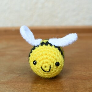 May include: A crocheted bee toy with black and yellow stripes, white wings, and black eyes. The bee has a smiling face.