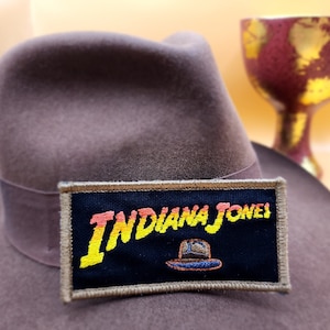 May include: A brown fedora hat with a black and beige embroidered patch that says "Indiana Jones" in yellow and orange letters. The patch also features a small blue and white fedora hat graphic.