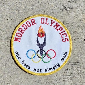 Pode incluir: Remendo bordado circular com as palavras "MORDOR OLYMPICS" em vermelho, uma tocha com um olho e os anéis olímpicos. A frase "one does not simply walk" também está incluída. O remendo tem uma borda amarela.