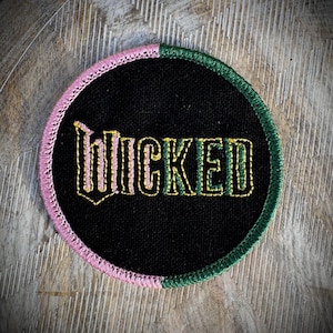 May include: A round black patch with pink and green stitching around the edges. The word "WICKED" is embroidered in yellow thread on the patch.