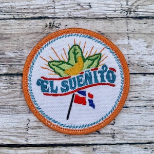 Puede incluir: Un parche blanco bordado con borde naranja. El parche presenta una hoja verde con un centro amarillo y una bandera roja, blanca y azul. El texto "EL SUEÑITO" está escrito en una pancarta azul.