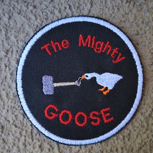 May include: A black and white embroidered patch with a white goose holding a hammer. The text "The Mighty Goose" is embroidered in red on the patch.