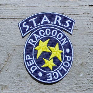 May include: A navy blue embroidered patch with white text and a white border. The patch features the text "S.T.A.R.S." above a circular design with "RACCOON POLICE DEP" and three yellow stars.