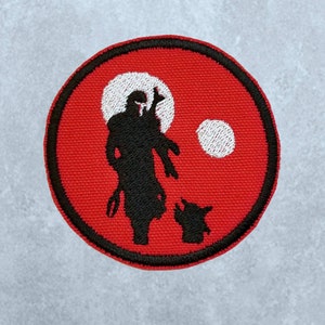 May include: A red circular embroidered patch featuring a black silhouette of a figure holding a weapon and a smaller figure standing beside it. The background has two white circles.