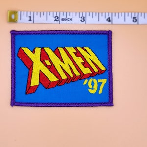Może przedstawiać: Naszywka haftowana w kolorze niebieskim i ż&oacute;łtym z tekstem "X-MEN '97" w stylu 3D.