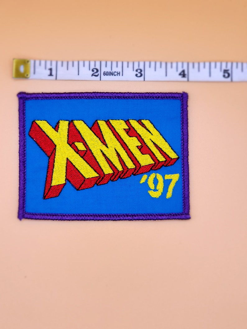 Może przedstawiać: Naszywka haftowana w kolorze niebieskim i ż&oacute;łtym z tekstem "X-MEN '97" w stylu 3D.