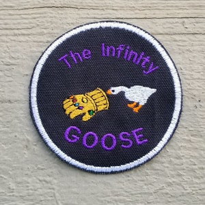 May include: A black and white circular patch with a white border. The patch features a purple embroidered design that says "The Infinity Goose" with a white goose biting a gold gauntlet with purple gems.
