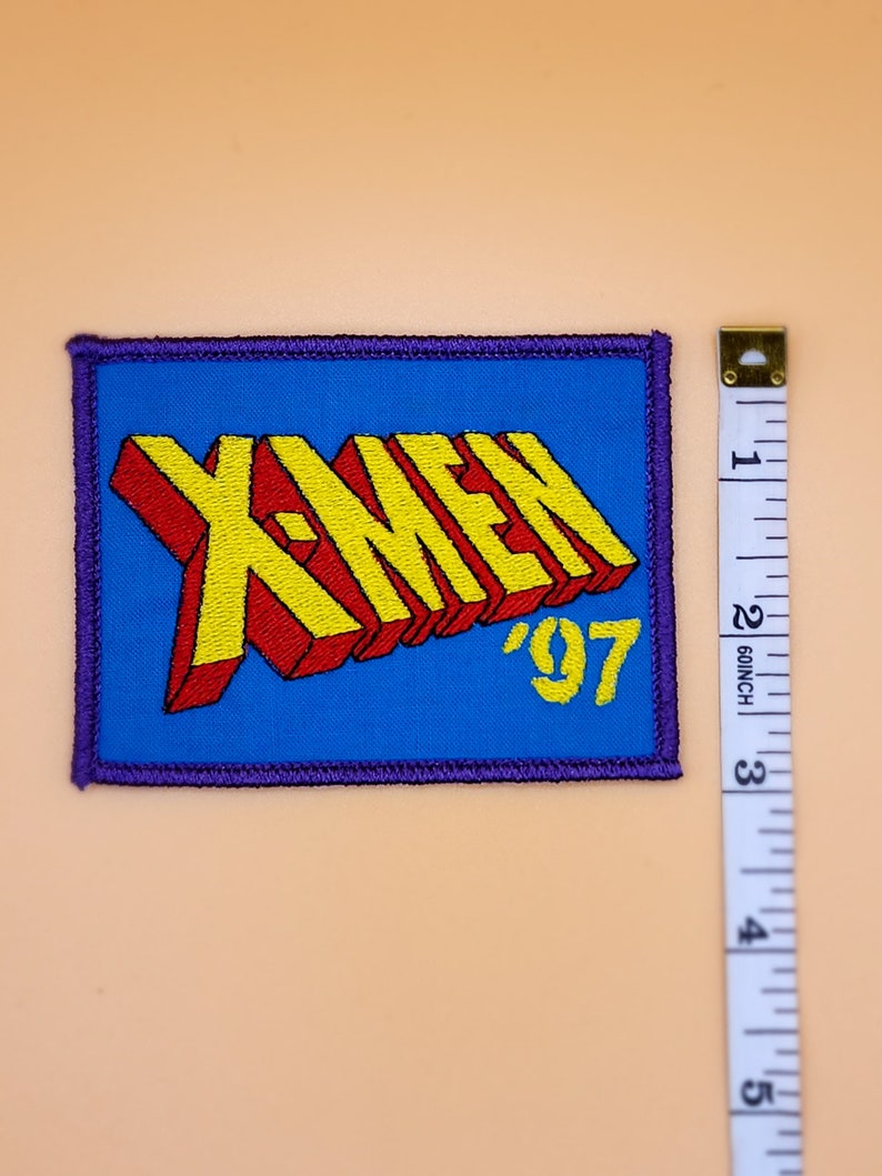 Może przedstawiać: Niebieska naszywka haftowana z napisem "X-Men '97" w ż&oacute;łtych i czerwonych literach.
