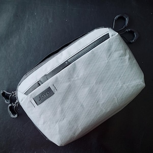 May include: A white, zippered pouch with a black J.S.G. logo and a black zipper pull. The pouch has a diamond pattern and is made of a lightweight, durable material.