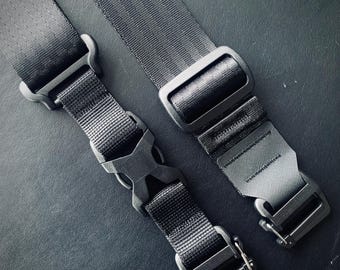 Op de afbeelding: Twee zwarte verstelbare banden met metalen gespen. Een band heeft een zwarte plastic clip. De banden liggen op een donkergrijs oppervlak.