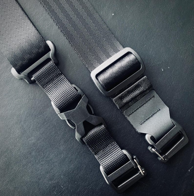 May include: Two black adjustable straps with metal buckles. One strap has a black plastic clip. The straps are on a dark gray surface.