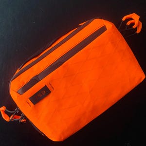 May include: Bright orange rectangular bag with a diamond pattern. The bag has black zippers and a small black tag with white text. Orange loops are attached to the bag with black cords.