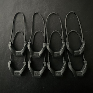 Puede incluir: Ocho tiradores de cremallera negros con forma hexagonal y el texto "JSD" impreso en cada uno.