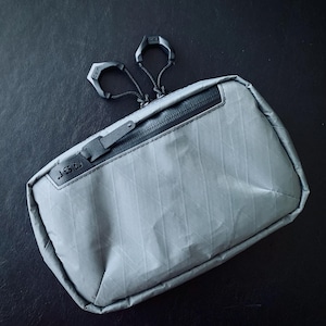 May include: A gray zippered pouch with a black zipper and a black pull tab. The pouch has a textured fabric and the text "JST" is printed on the front.