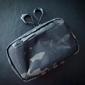 May include: A black and grey camouflage zippered pouch with a black zipper pull and a black J.S.D. logo.