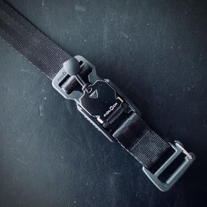 Puede incluir: Hebilla Fidlock negra con una correa de nailon negra. La hebilla tiene el logotipo de Fidlock y un cierre magnético. La correa es tejida y tiene un lazo de metal en el extremo. La hebilla es de plástico y metal.