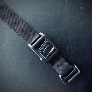 May include: A close-up shot of a black Fidlock buckle and strap. The buckle is rectangular with the brand name printed on it. The strap is made of a woven material. The background is a dark, textured surface.