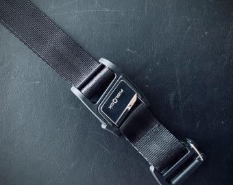 Op de afbeelding: Close-up van een zwarte Fidlock-gesp en -riem. De gesp is rechthoekig met de merknaam erop gedrukt. De riem is gemaakt van een geweven materiaal. De achtergrond is een donker, getextureerd oppervlak.