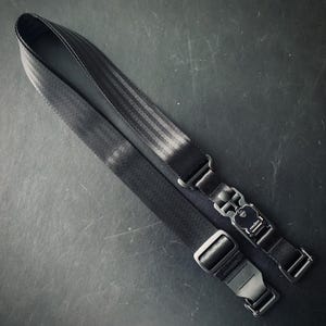 May include: Black camera strap with a seatbelt-style design. The strap features a quick-release buckle and adjustable length. The strap is made of durable, woven material and is designed for secure camera carrying.