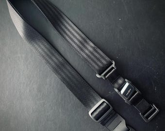 Op de afbeelding: Zwarte camerariem met een gestreept patroon en metalen en plastic gespen. De riem is ontworpen om verstelbaar en veilig te zijn, met een duurzame constructie. De riem is ongeveer 127 cm lang.
