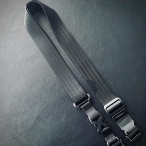 May include: A black camera strap with two adjustable buckles. The strap is made of a durable, woven material.