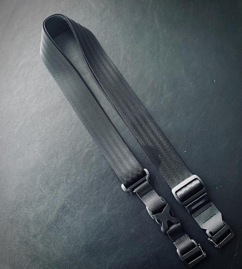 May include: A black camera strap with two adjustable buckles. The strap is made of a durable, woven material.