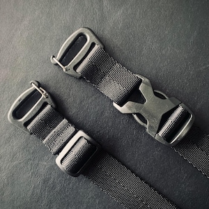 Op de afbeelding: Close-up van twee zwarte nylon singelbanden met metalen hardware. Eén band heeft een zijgesp, terwijl de andere een metalen haak heeft. De banden zijn geplaatst tegen een donkere achtergrond, waardoor de textuur en details worden benadrukt.