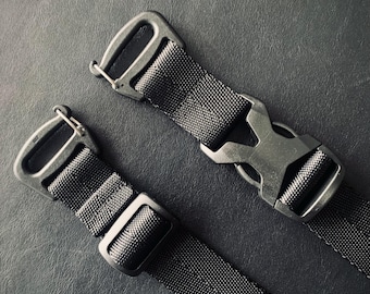 Op de afbeelding: Close-up van twee zwarte nylon singelbanden met metalen hardware. Eén band heeft een zijgesp, terwijl de andere een metalen haak heeft. De banden zijn geplaatst tegen een donkere achtergrond, waardoor de textuur en details worden benadrukt.