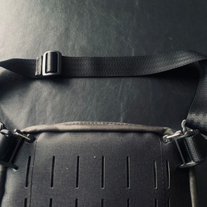 May include: A black crossbody bag with a black adjustable strap and silver buckles. The bag has a textured front panel with multiple rows of small holes.