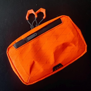 May include: Orange zippered pouch with a black zipper and a black logo that says "1SD". The pouch has a diamond pattern and two orange pull tabs.