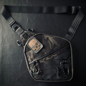May include: A black tactical sling bag with a camouflage pattern and a skull-shaped patch. The bag has a zippered pocket and a buckle closure.