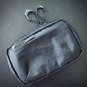 May include: A black zippered pouch with a JSD logo and two black clips attached to the zipper pull.