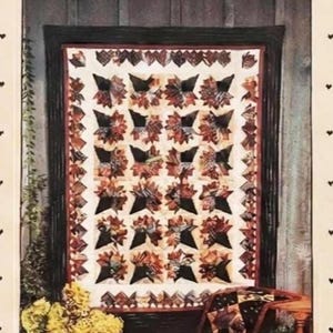 May include: A rectangular paper pattern envelope titled Bride's Bouquet #158 by Country Threads. The cover features a photograph of a quilt with a leaf pattern in brown, cream, and black, displayed on a wooden wall with yellow flowers nearby.