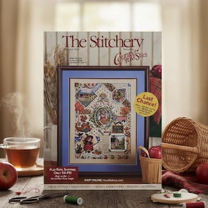 May include: A magazine titled "The Stitchery" featuring counted cross stitch designs. The cover art is a framed cross-stitch sampler with various autumn-themed motifs. A cup of tea, apples, and sewing supplies are on a wooden table.