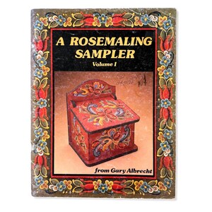 May include: A softcover book titled A ROSEMALING SAMPLER Volume I from Gary Albrecht. The cover features a decorative border of painted blue and red flowers and a central image of a red wooden box with intricate floral rosemaling patterns.