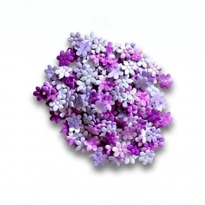 May include: A collection of small, five-petaled fabric flowers in various shades of purple, ranging from light lavender to deep violet. The flowers are arranged in a dense, circular cluster, each featuring a smooth, slightly shiny texture.