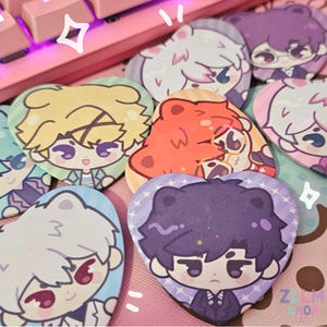 Könnte beinhalten: Ein Set aus neun herzförmigen Knöpfen mit Anime-Charakteren mit verschiedenen Haarfarben und Kleidung. Die Knöpfe befinden sich auf einer rosa Oberfläche mit einer Tastatur im Hintergrund.