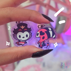May include: Two glittery acrylic keychains featuring cartoon characters. One keychain has a purple and white character with a crown and cape. The other keychain has a red, black, and white character with heart-shaped markings.