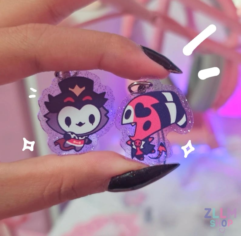 May include: Two glittery acrylic keychains featuring cartoon characters. One keychain has a purple and white character with a crown and cape. The other keychain has a red, black, and white character with heart-shaped markings.