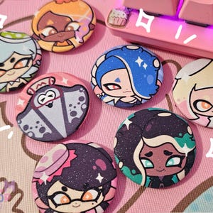 Puede incluir: Ocho botones redondos de colores con personajes del videojuego Splatoon. Los botones presentan diferentes personajes con diversas expresiones y poses. Los botones están decorados con purpurina y tienen un acabado brillante.