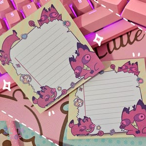 Könnte beinhalten: Zwei quadratische Haftnotizen mit hellgelbem Hintergrund und rosa Rand mit Cartoon-Monstern. Die Notizen haben liniertes Papier und sind von rosa und lila Tastaturtasten und einer rosa Schreibtischunterlage umgeben.