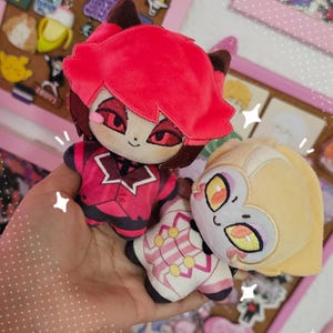 Puede incluir: Dos muñecos de peluche de personajes de dibujos animados. Uno tiene el pelo rojo y un atuendo rojo, el otro tiene el pelo rubio y un atuendo blanco y rosa. Los muñecos se sostienen en una mano, con un fondo de alfileres y pegatinas.